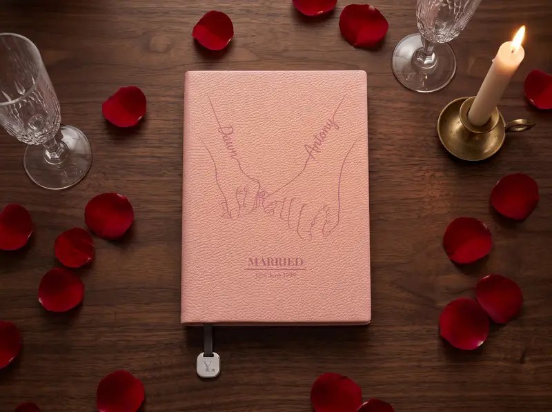 A Blush notebook engraved with two hands, little fingers linked and a name lettered along each forearm, above the word Married and a date, on a dark walnut table strewn with deep red rose petals beside a lit candle.