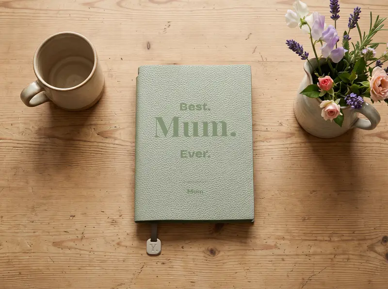 A Willow notebook engraved “Best. Mum. Ever.”, on a scrubbed kitchen table beside a stoneware mug and a jug of garden flowers.