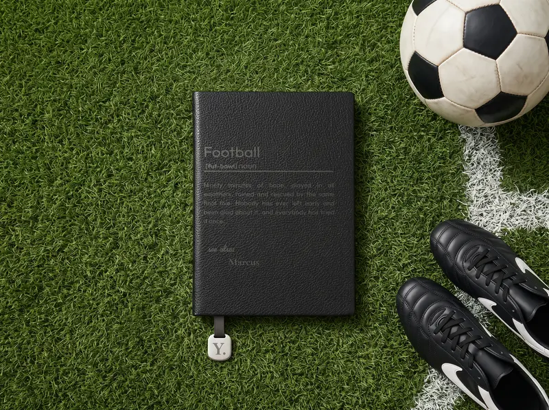 An Onyx notebook engraved with a football definition, on the grass at the edge of a pitch beside a ball and a pair of boots.