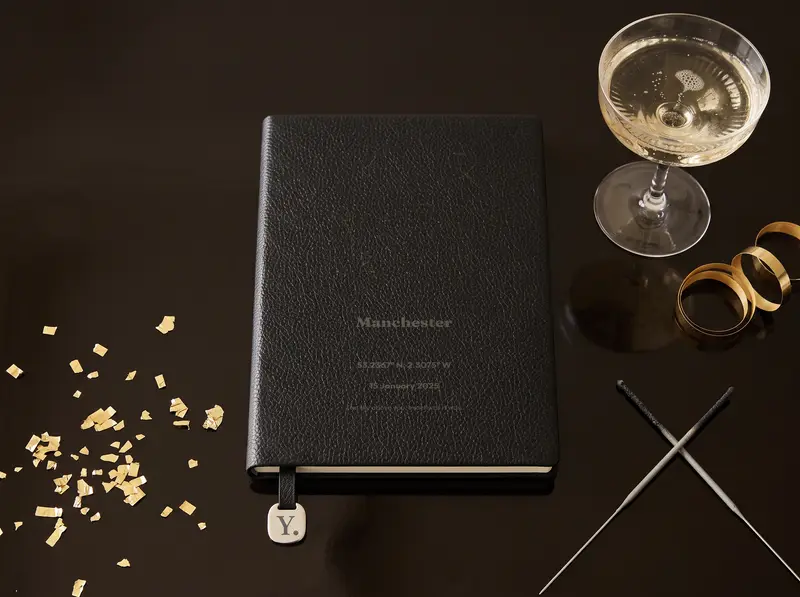 A personalised, laser-engraved A5 notebook on a dark table lit by gold confetti, beside a coupe glass and a pair of sparklers.