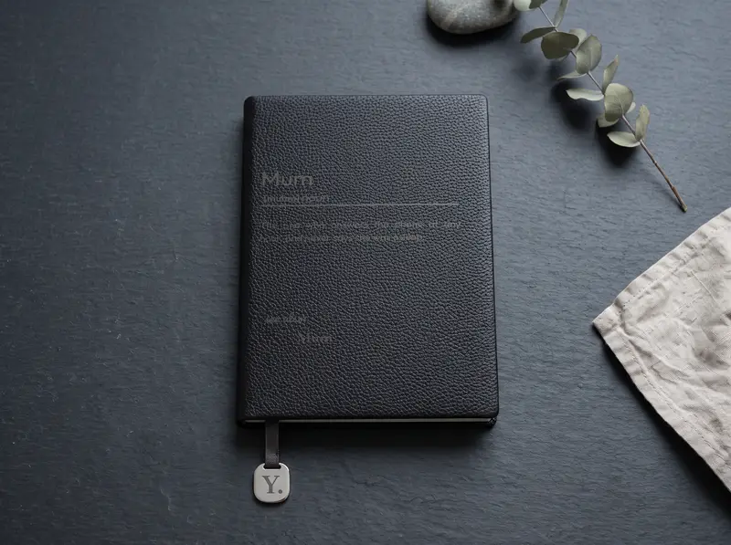 An Onyx notebook engraved with a dictionary entry for “Mum” and a definition written by the buyer, on dark slate lit from one side.