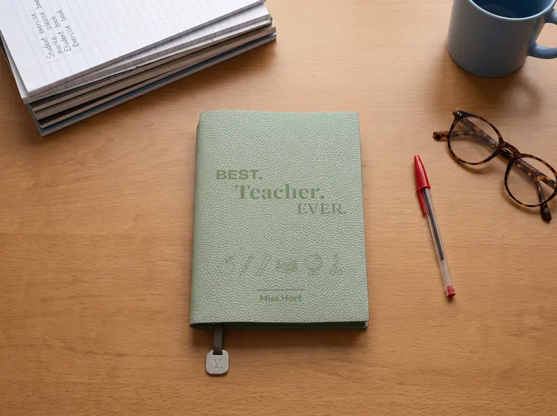 A Willow notebook engraved “Best. Teacher. Ever.”, on a desk beside marked exercise books, a red pen and reading glasses.
