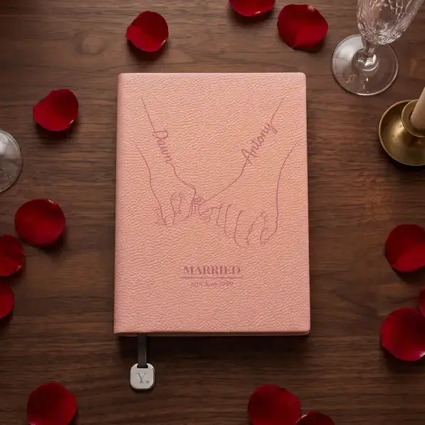A personalised, laser-engraved A5 notebook on a dark walnut table strewn with deep red rose petals, beside two champagne flutes and a lit candle.