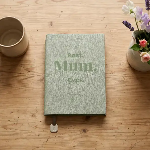 A personalised, laser-engraved A5 notebook on a scrubbed kitchen table, beside a stoneware mug and a jug of garden flowers.