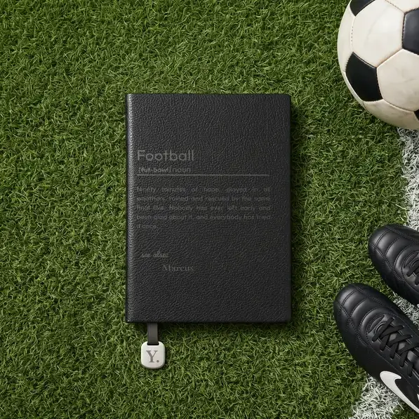 A personalised, laser-engraved A5 notebook on the grass at the edge of a pitch, beside a ball and a pair of boots.