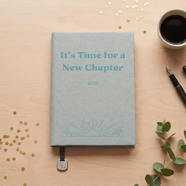 A personalised, laser-engraved A5 notebook on a pale oak table on New Year's morning, beside black coffee, a fountain pen and a scatter of gold foil stars.
