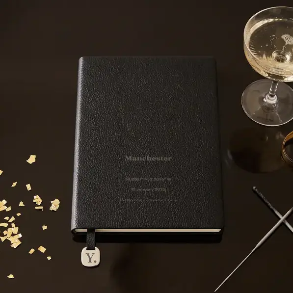 A personalised, laser-engraved A5 notebook on a dark table lit by gold confetti, beside a coupe glass and a pair of sparklers.