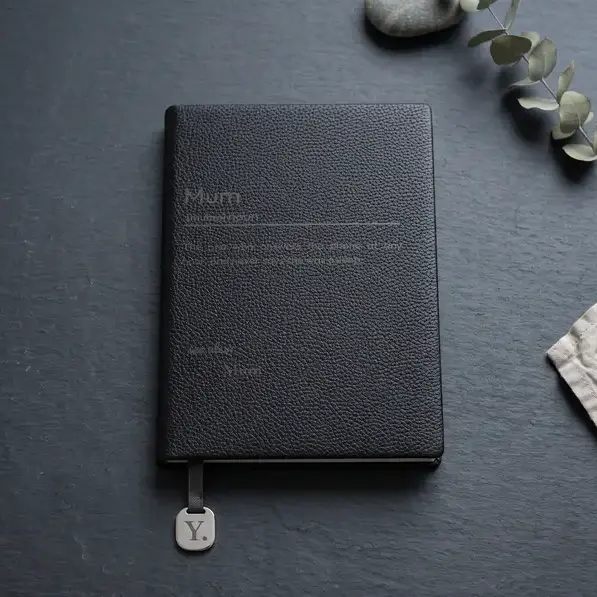 A personalised, laser-engraved A5 notebook on dark slate lit from one side, beside a sprig of eucalyptus and folded linen.