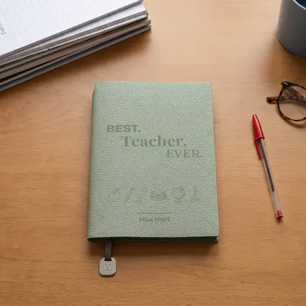 A personalised, laser-engraved A5 notebook on a teacher's desk, beside marked exercise books, a red pen and reading glasses.